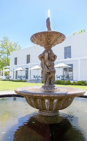 Une fontaine en pierre ornée d’une statue, entourée d’un bassin d’eau. Au fond, on aperçoit un bâtiment blanc doté d’une terrasse et de parasols.