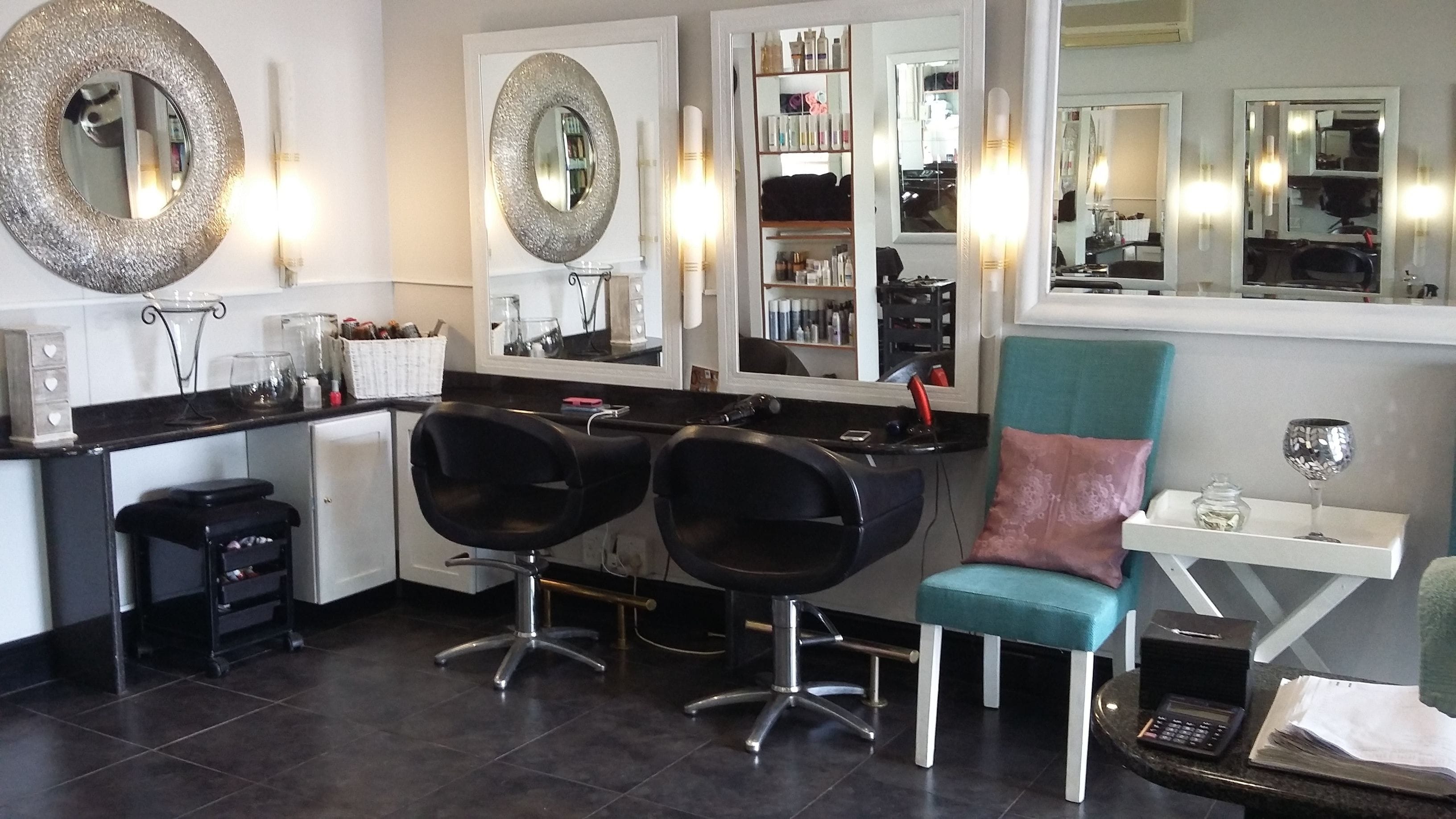 Un salon avec deux fauteuils noirs, un fauteuil turquoise et un comptoir blanc agrémenté d’un miroir et d’étagères remplies de produits.