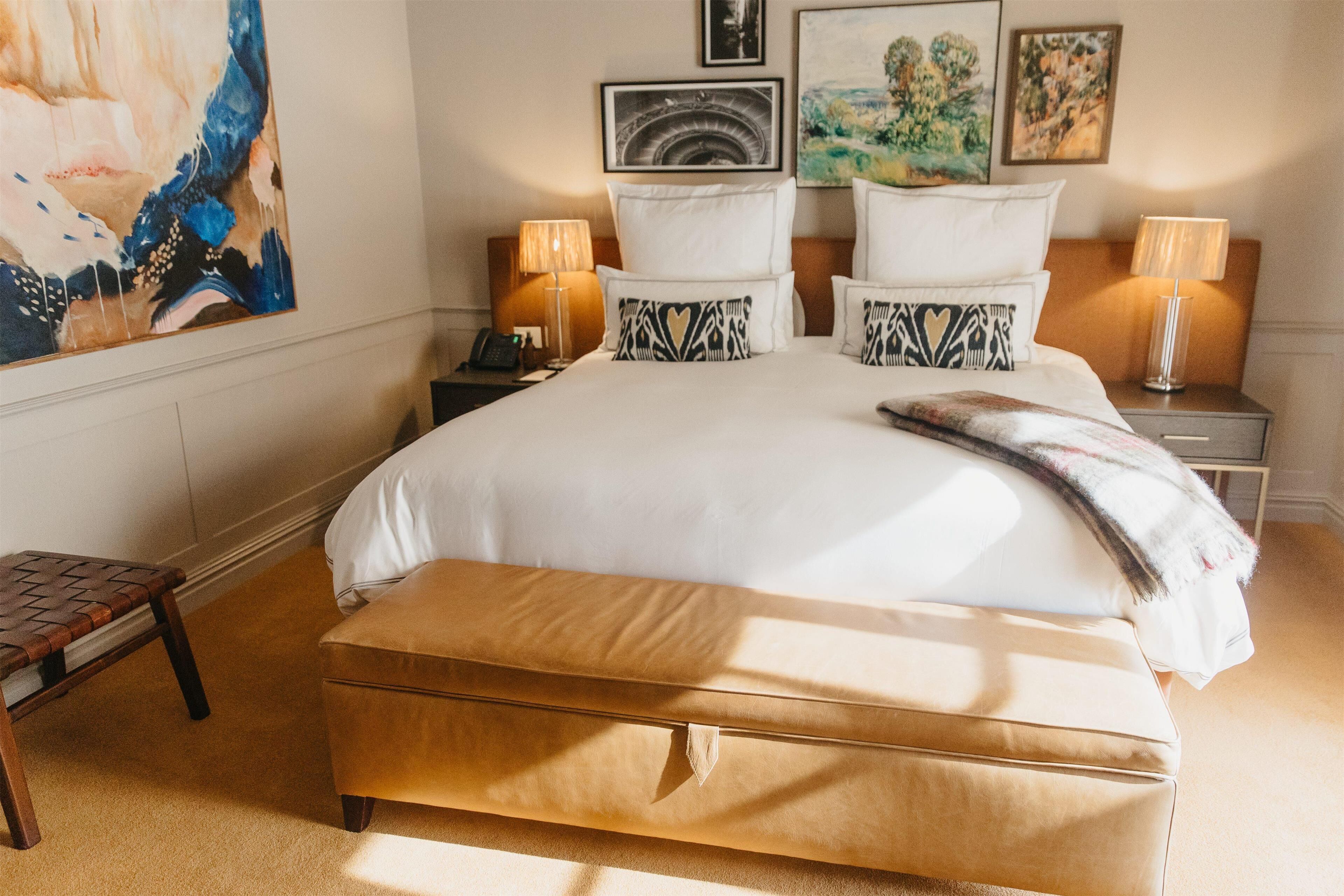 Un cuarto de hotel ordenado con una cama grande, dos mesitas de noche con lámparas, un banco y obras de arte en las paredes.