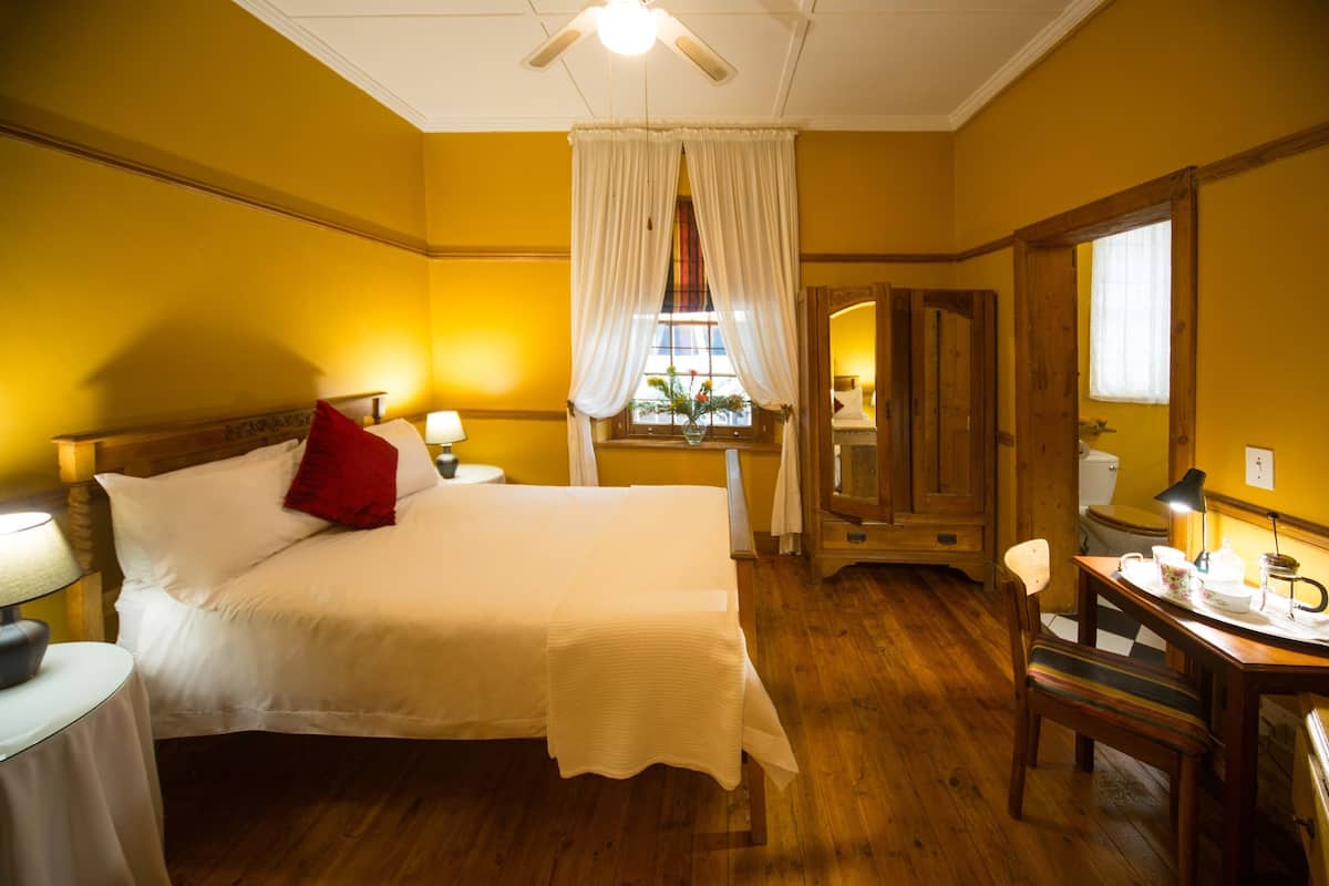 A hotel room with a bed, a desk with a chair, a window with curtains, and a wooden floor.