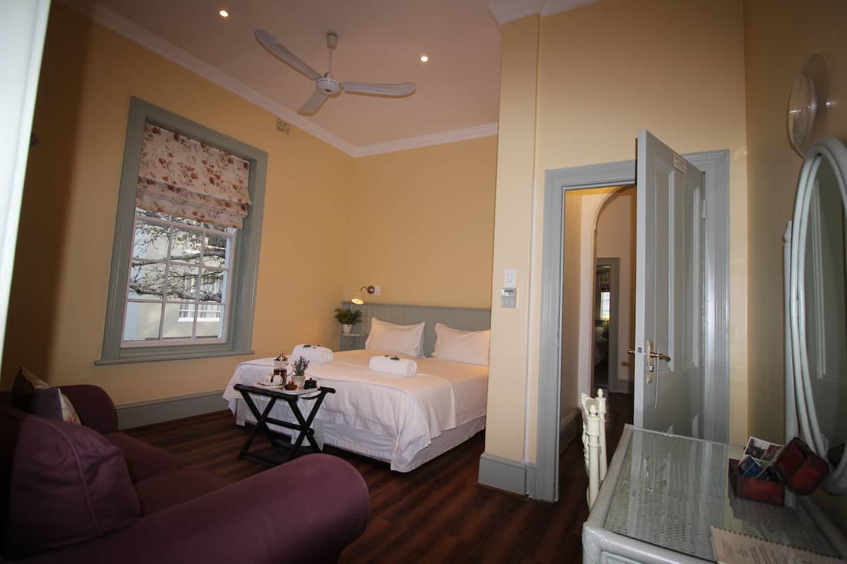 A bedroom with a bed, a ceiling fan, a window with a floral blind, a small table with a plant, and a door leading to another room.