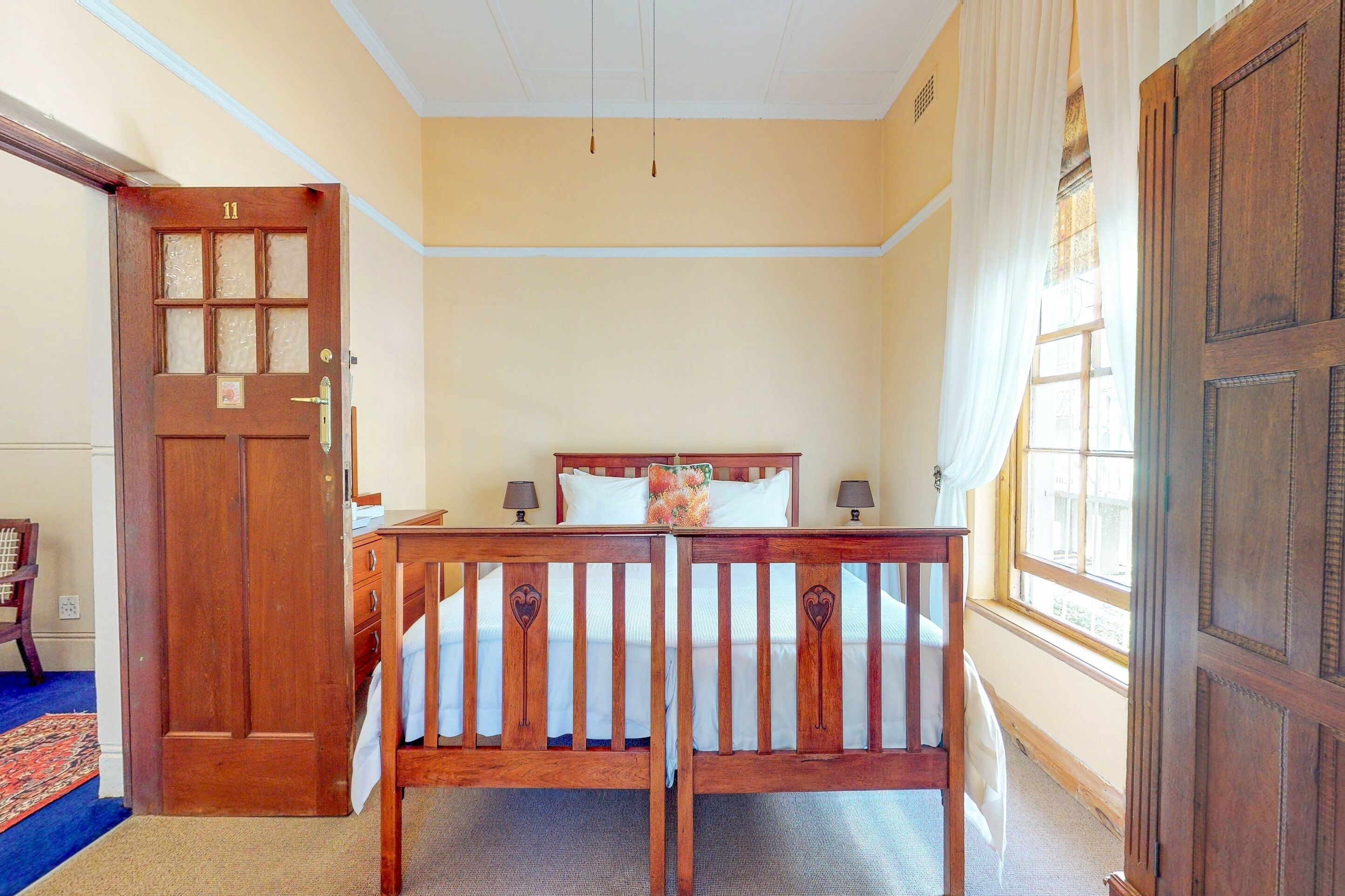 Ein Zimmer mit einem Holzbett, einer Tür mit einer Nummer und einem mit Vorhängen versehenen Fenster.