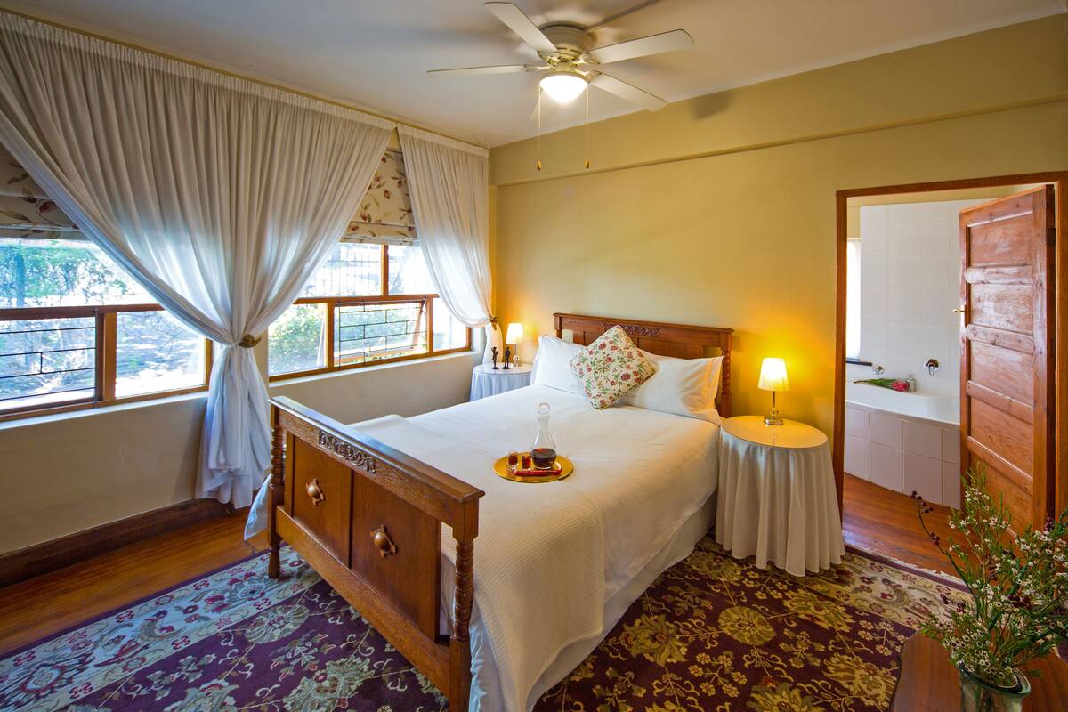 A bedroom with a wooden bed, a nightstand with a lamp, a window with curtains, and a view of the outside.