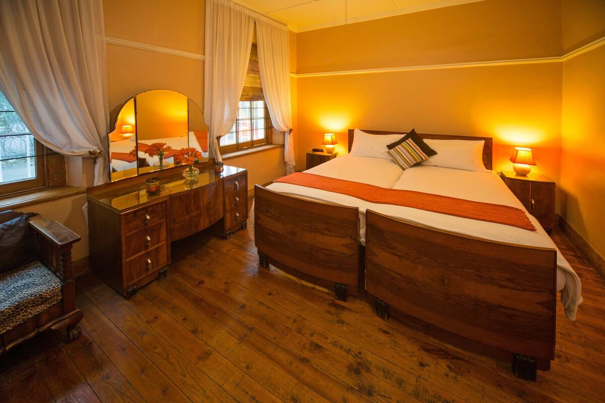 A double bed with a wooden headboard, two bedside tables with lamps, a wooden dresser, a mirror, and a window with curtains.