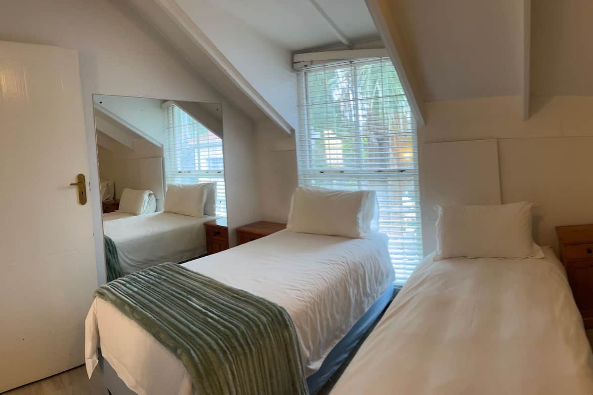 A hotel room with two single beds, a small bedside table with a lamp, a mirror, and a window with blinds.
