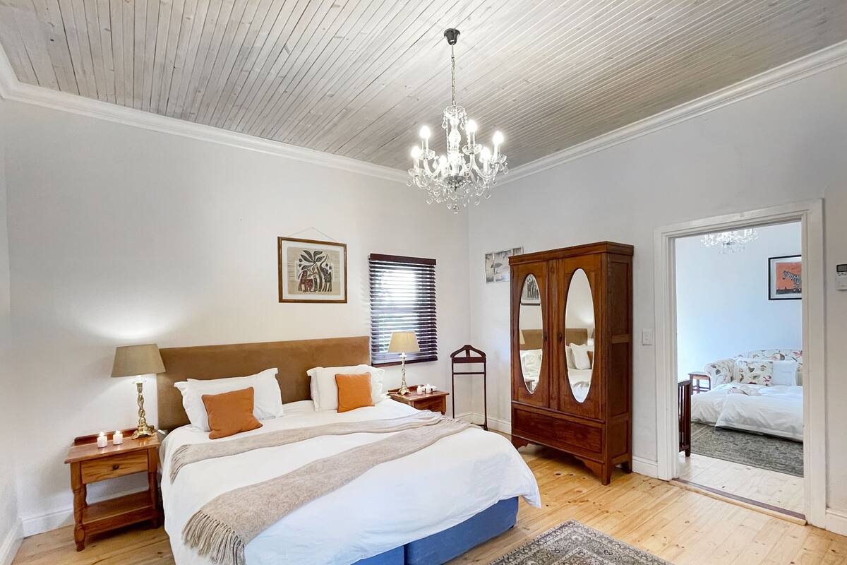 A bedroom with a large bed, a wooden wardrobe, a chandelier, and a view into another room.