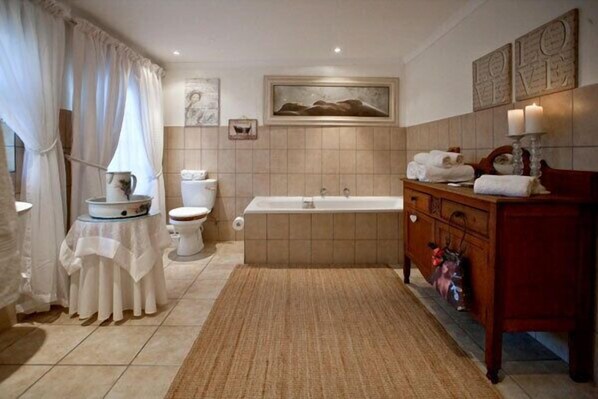 Un baño con bañera, inodoro y lavabo, con piso de baldosas color beige y tocador de madera.