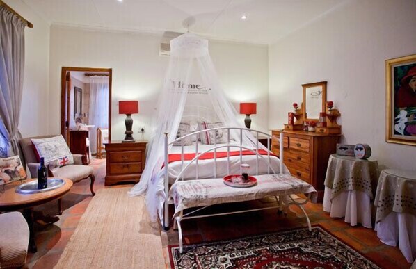 A bedroom with a canopy bed, a bedside table, a chair, a dresser, and a small table with a lamp. - Thylitshia Villa (Oudtshoorn)
