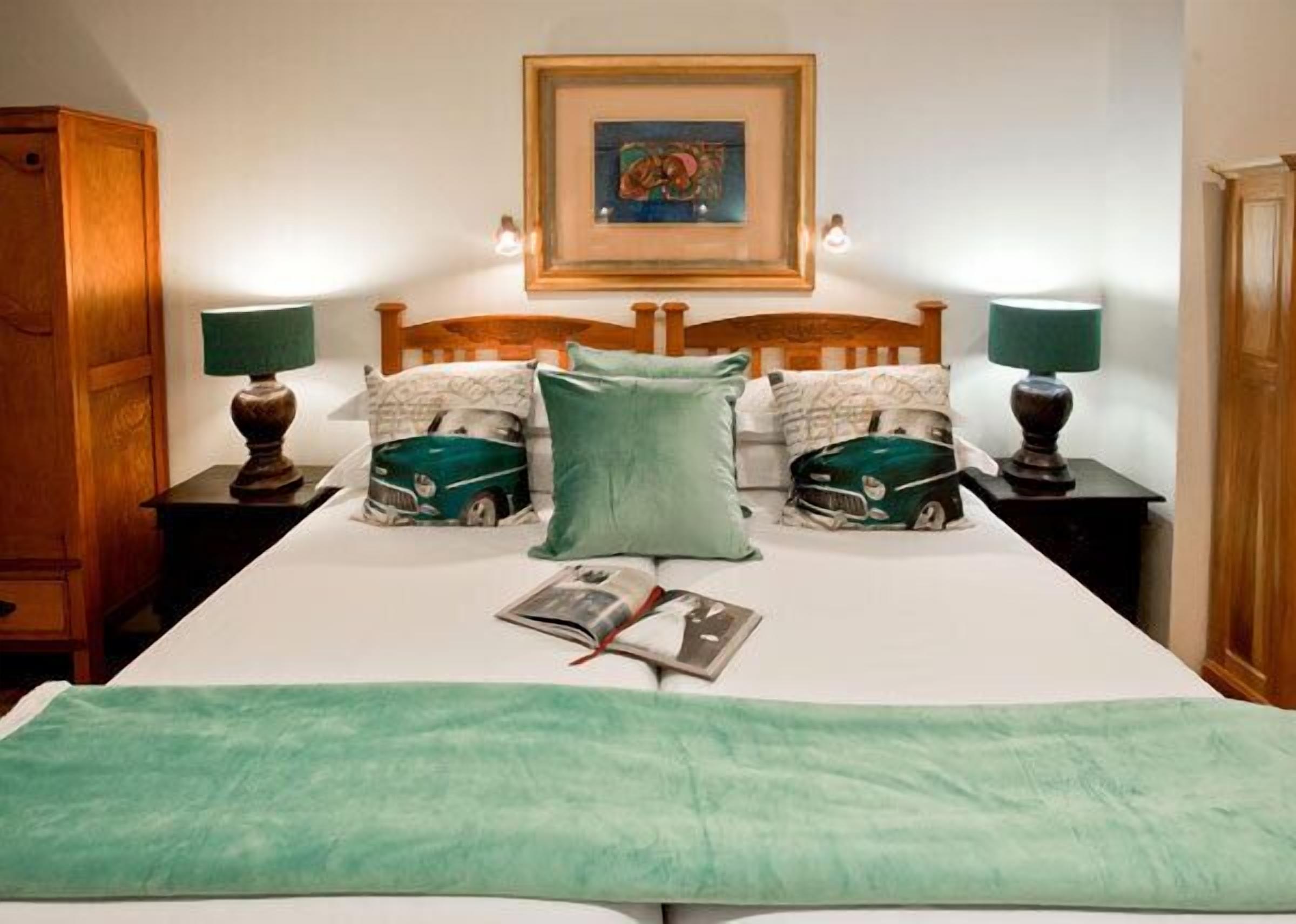 Una cama bien hecha con una manta verde, dos almohadas con estampados de autos, un cuadro enmarcado sobre la cama y dos lámparas de noche.