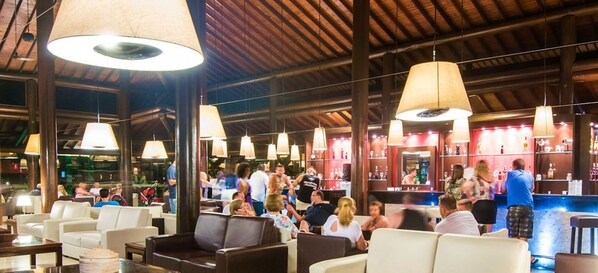 Zona de bar con varias lámparas colgantes, diferentes disposiciones de asientos y una barra con varios botellas.