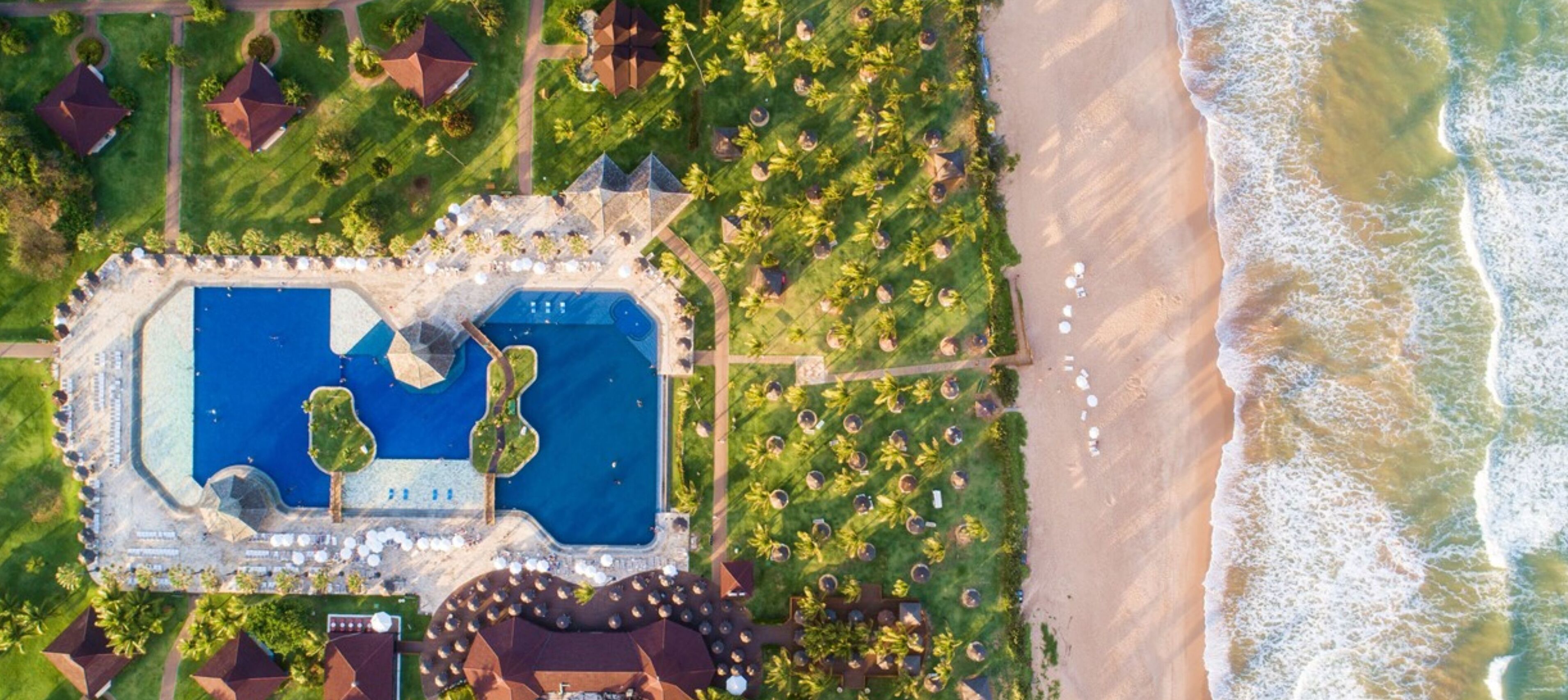 An aerial view of a resort with a large swimming pool, beach access, and lush landscaping.