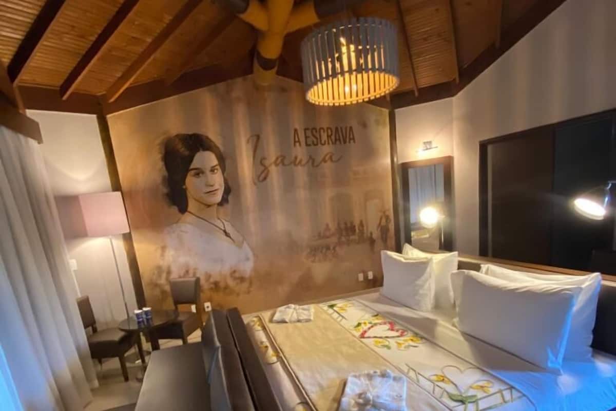 A bedroom with a bed, bedside tables, a desk, and a chair. There is a wall with a portrait and text.