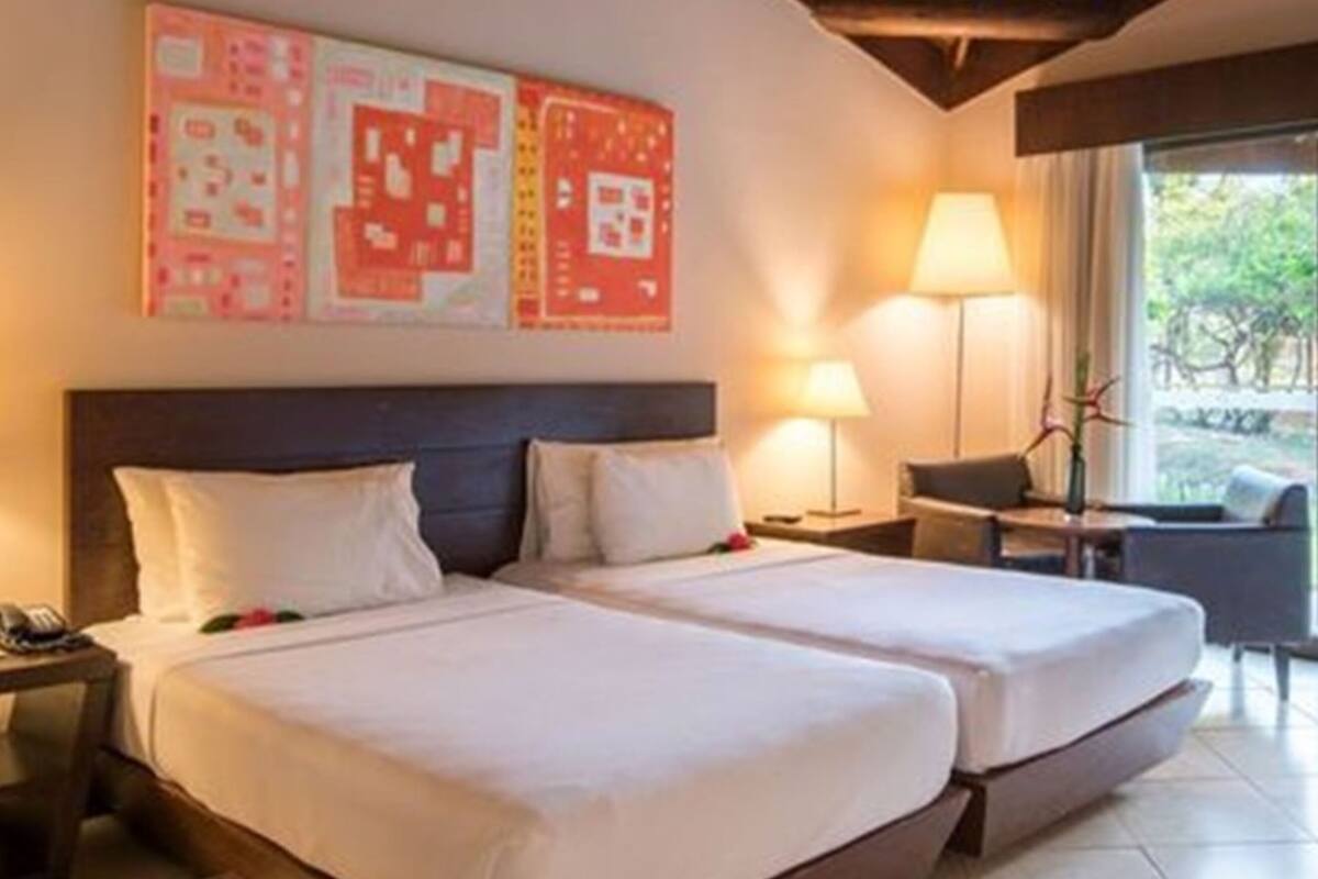 A hotel room with two beds, a desk, and a chair. There are two lamps on either side of the bed and a large window with a view of greenery outside.