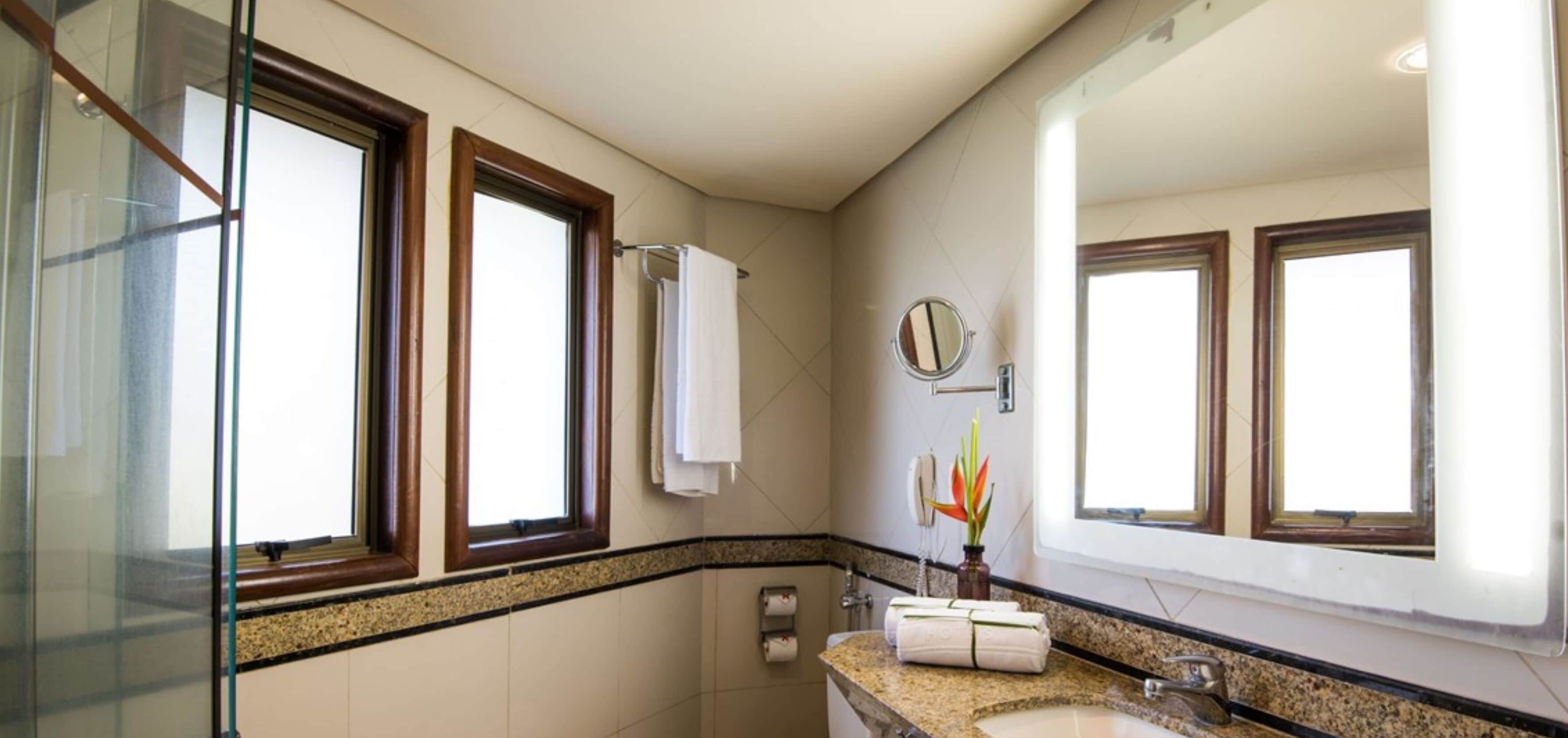 Un baño con cabina de ducha de vidrio, encimera de granito, lavabo, espejo y ventana con una toalla colgada en la pared.