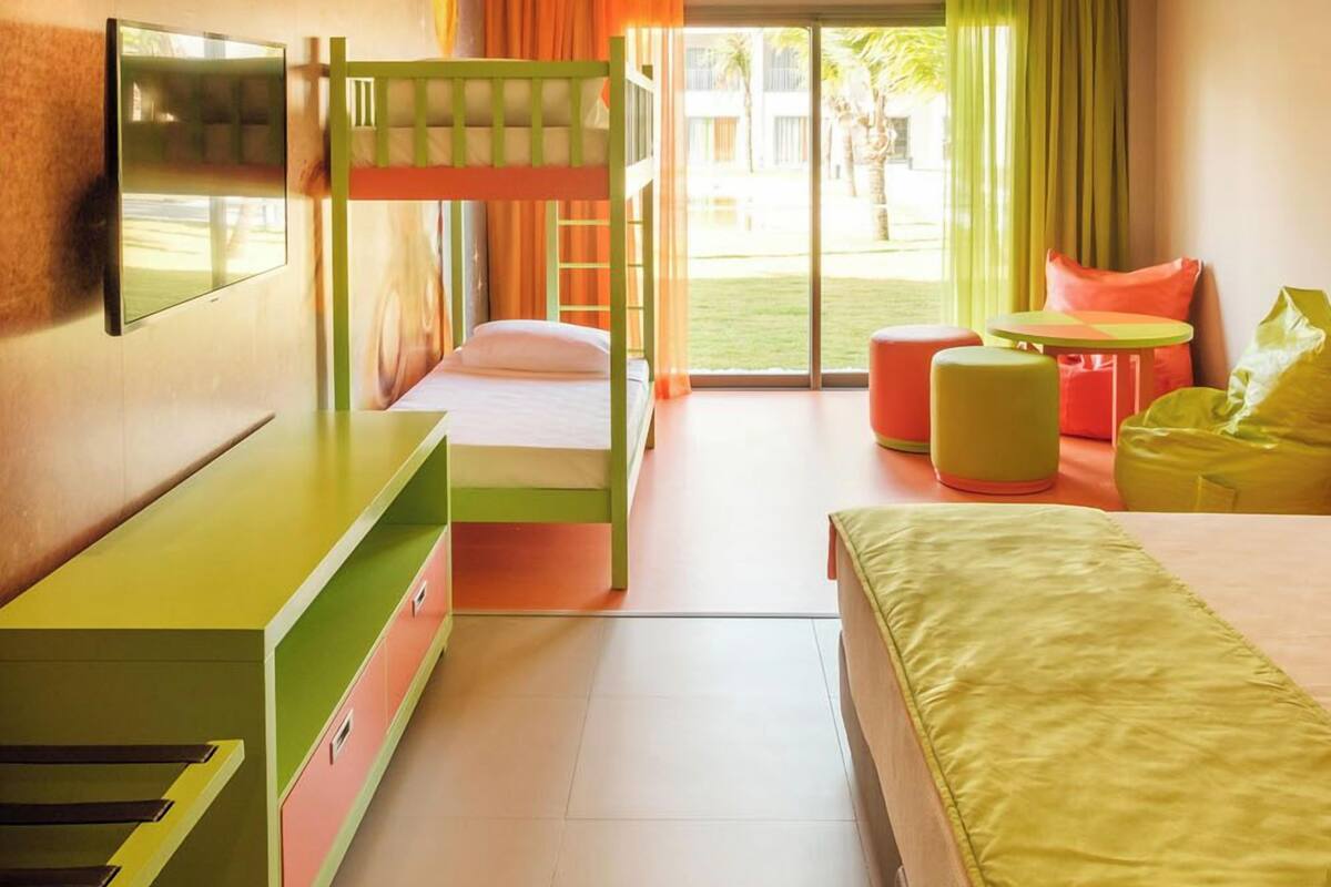 A brightly colored hotel room with bunk beds, a TV, a desk, and a window with a view of the outdoors.