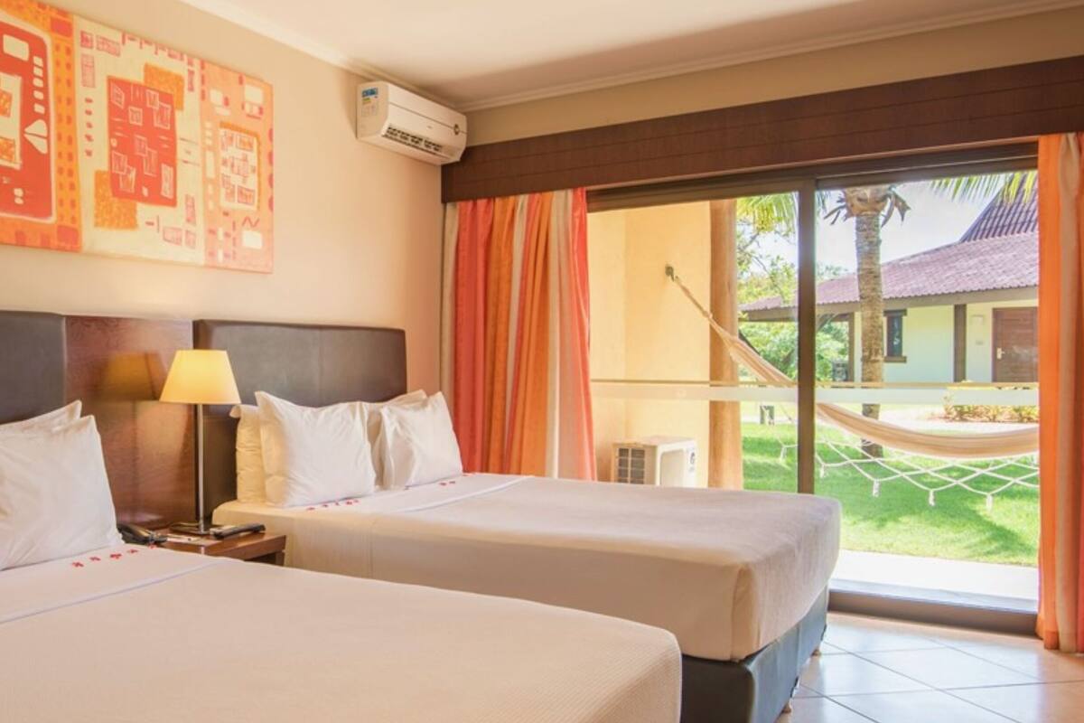 A hotel room with two beds, a nightstand, a lamp, and a large window with curtains.