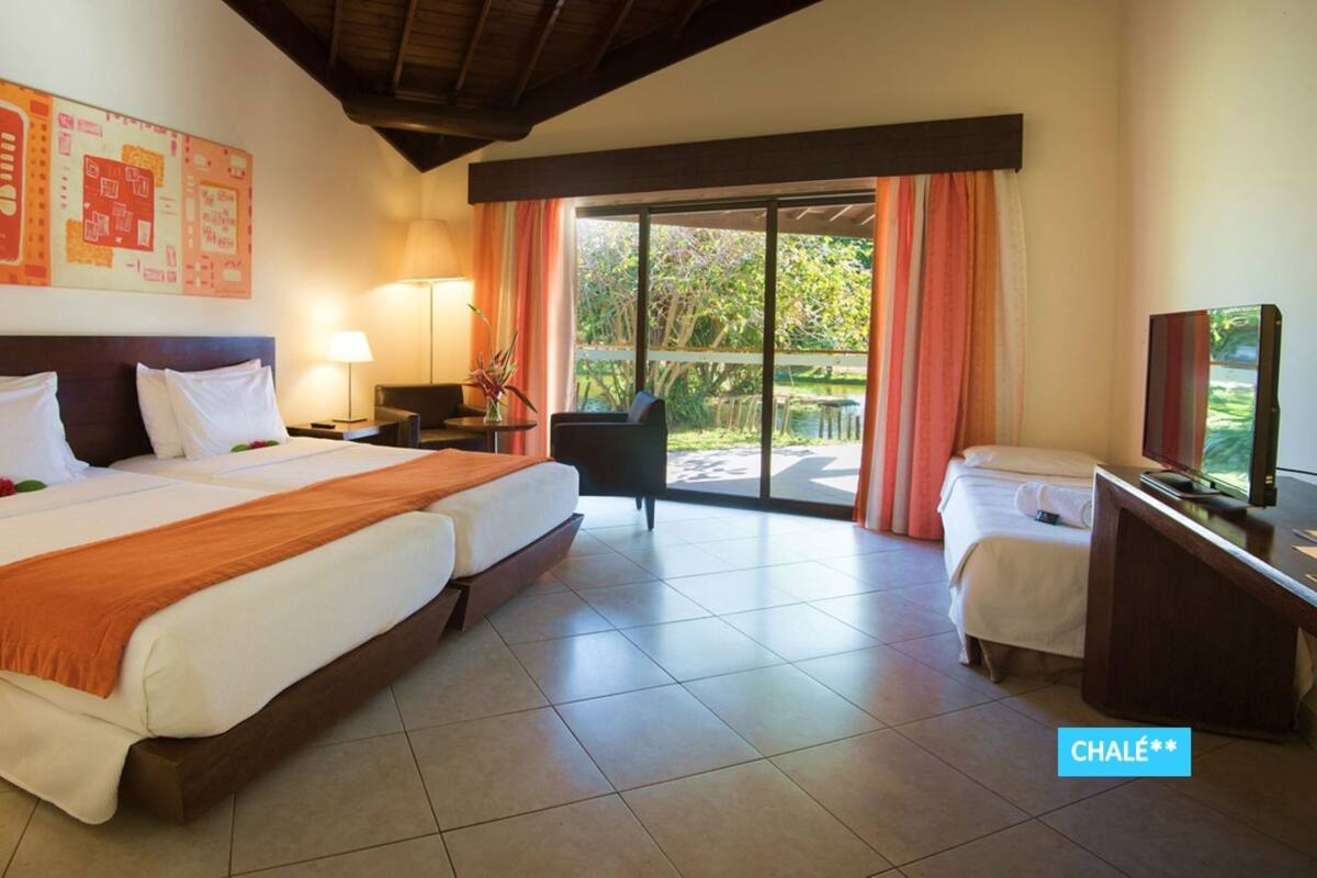 A hotel room with a large bed, a desk with a TV, a chair, and a balcony with curtains.