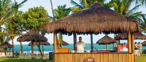 A beach bar with thatched umbrellas, a bartender, and lounge chairs.