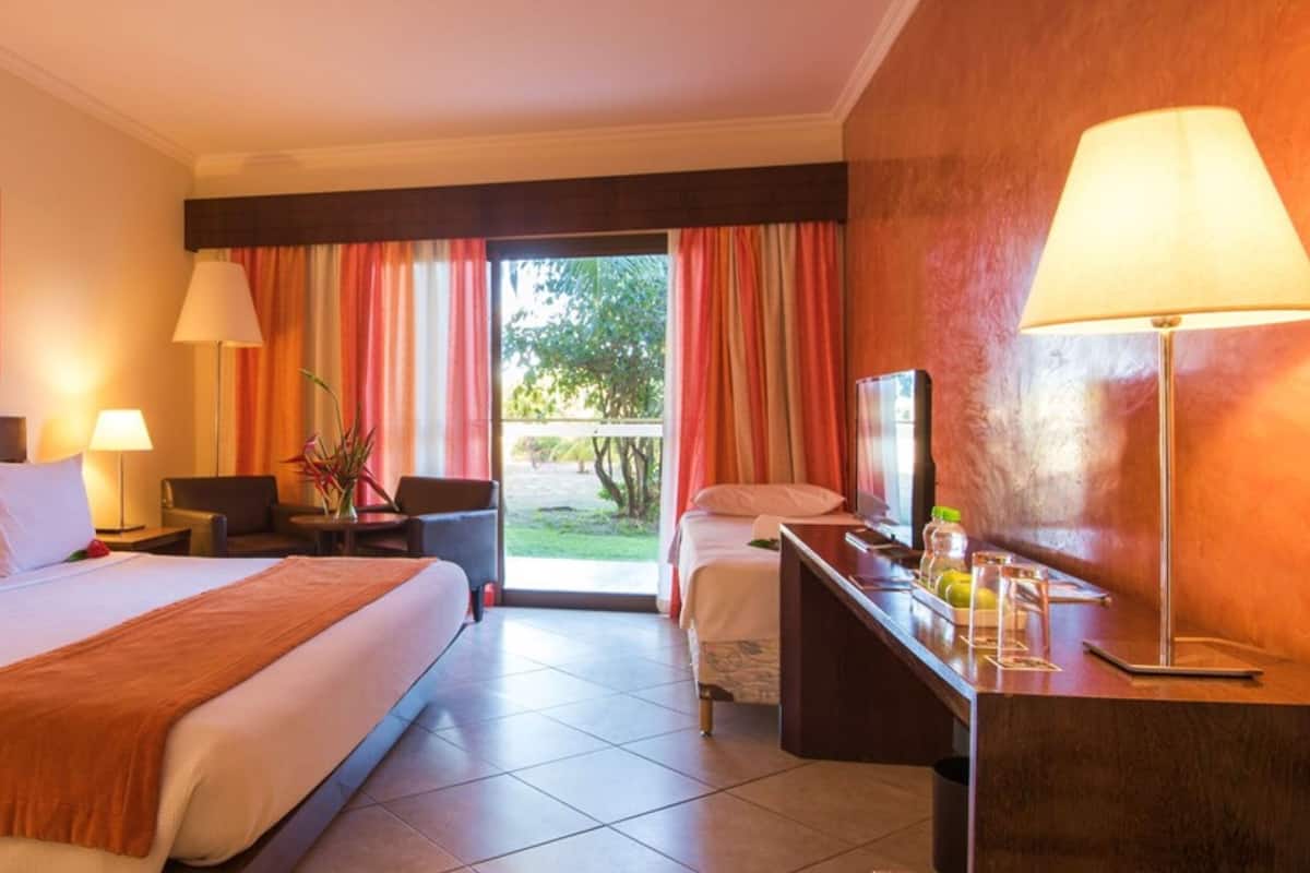 A hotel room with a large bed, a desk with a lamp, a chair, and a view of the outdoors through a sliding door.