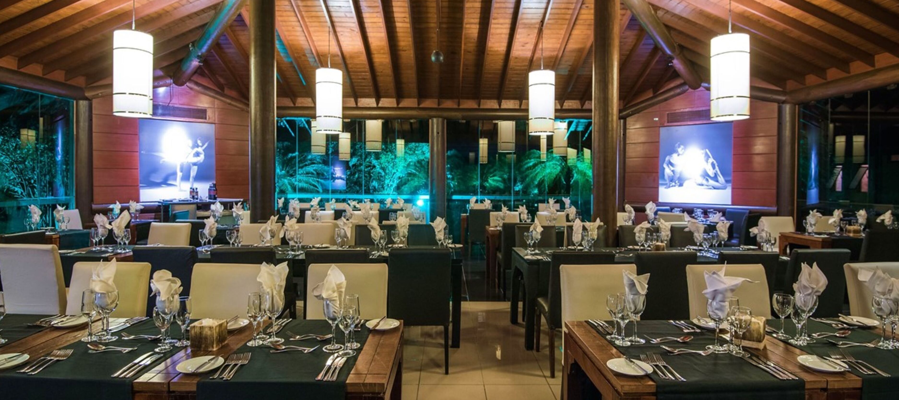 Een restaurant met meerdere tafels gedekt voor diners, met een houten plafond en grote ramen met uitzicht op groen.