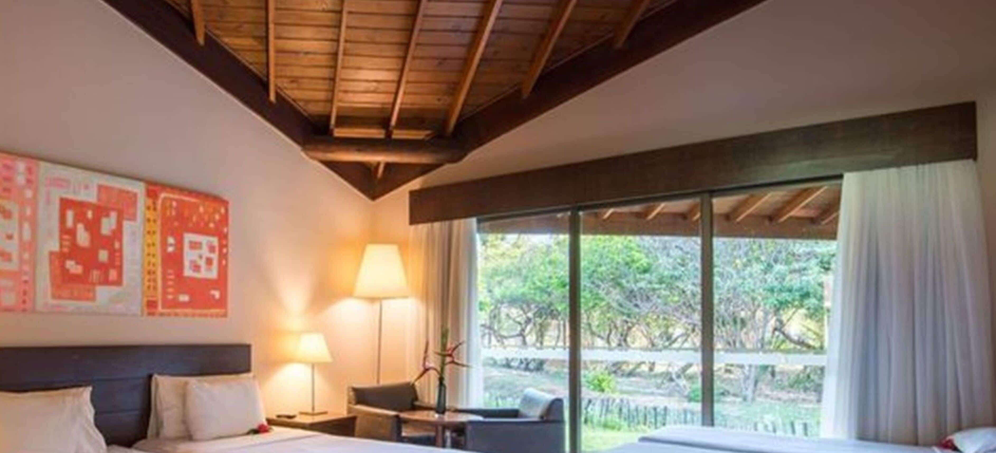 Habitación de hotel con cama, escritorio, silla y un amplio ventanal con vistas a árboles.