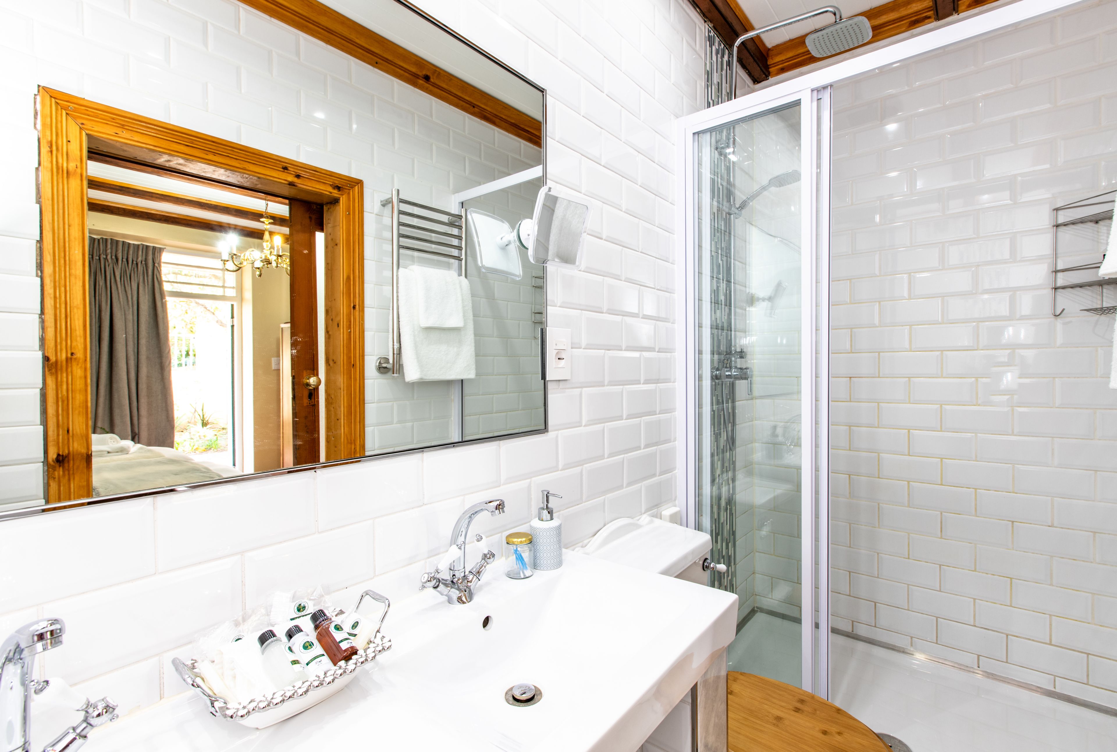 Ein Badezimmer mit weiß gefliesten Wänden, einem spiegelnden Holzrahmen, einem weißen Waschbecken mit Tabletts mit Toilettenartikeln und einer Duschkabine aus Glas.