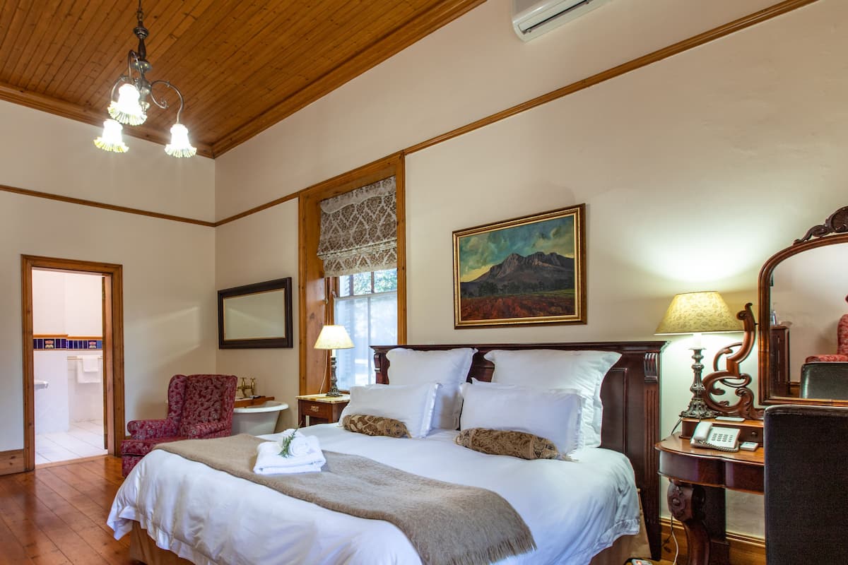 Ein Schlafzimmer mit einem großen Bett, Holzboden, einer Kandelaberleuchte, einem Gemälde, einem Sessel und einer Kommode.