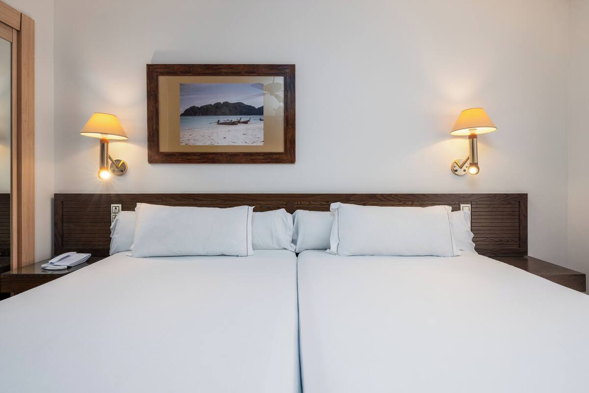 A double bed with white linens, two bedside lamps, a framed picture of a beach scene, and a wooden headboard.