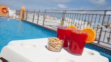 Escena junto a la piscina con dos bebidas, una con una rodaja de naranja, y un vaso de cacahuates sobre una silla blanca.