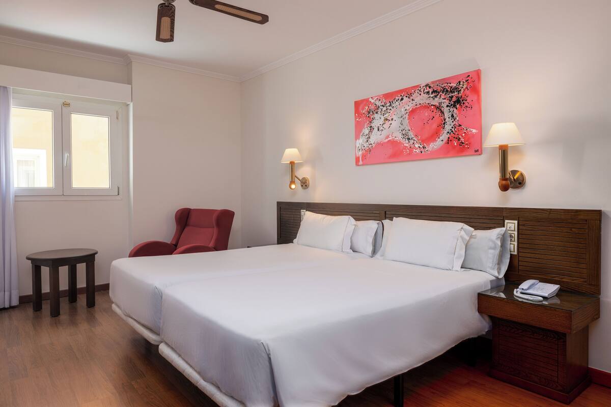 A hotel room with a large bed, two bedside lamps, a red chair, a small table, and a painting on the wall.