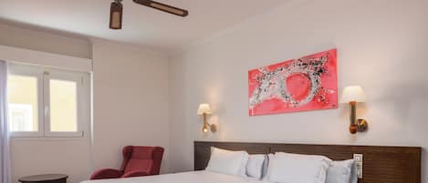 A hotel room with a large bed, two bedside lamps, a red chair, a small table, and a painting on the wall.