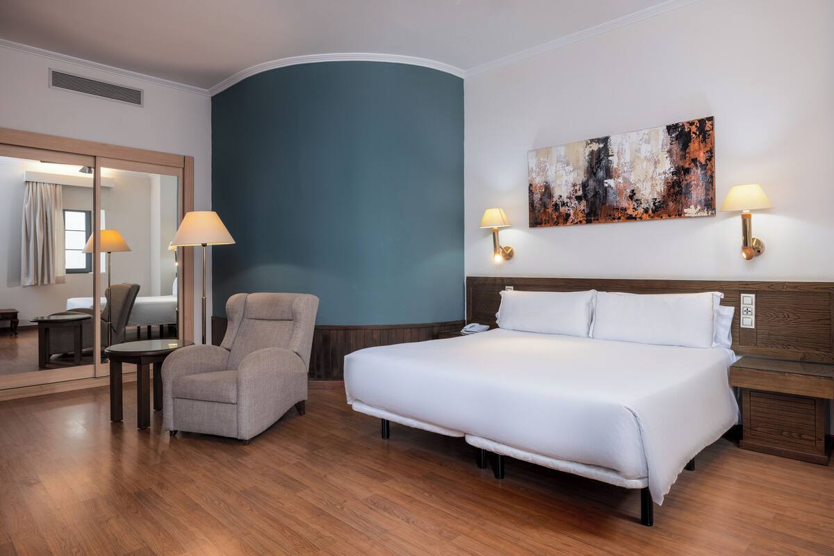 A hotel room with a large bed, a chair, a desk, and a painting on the wall.