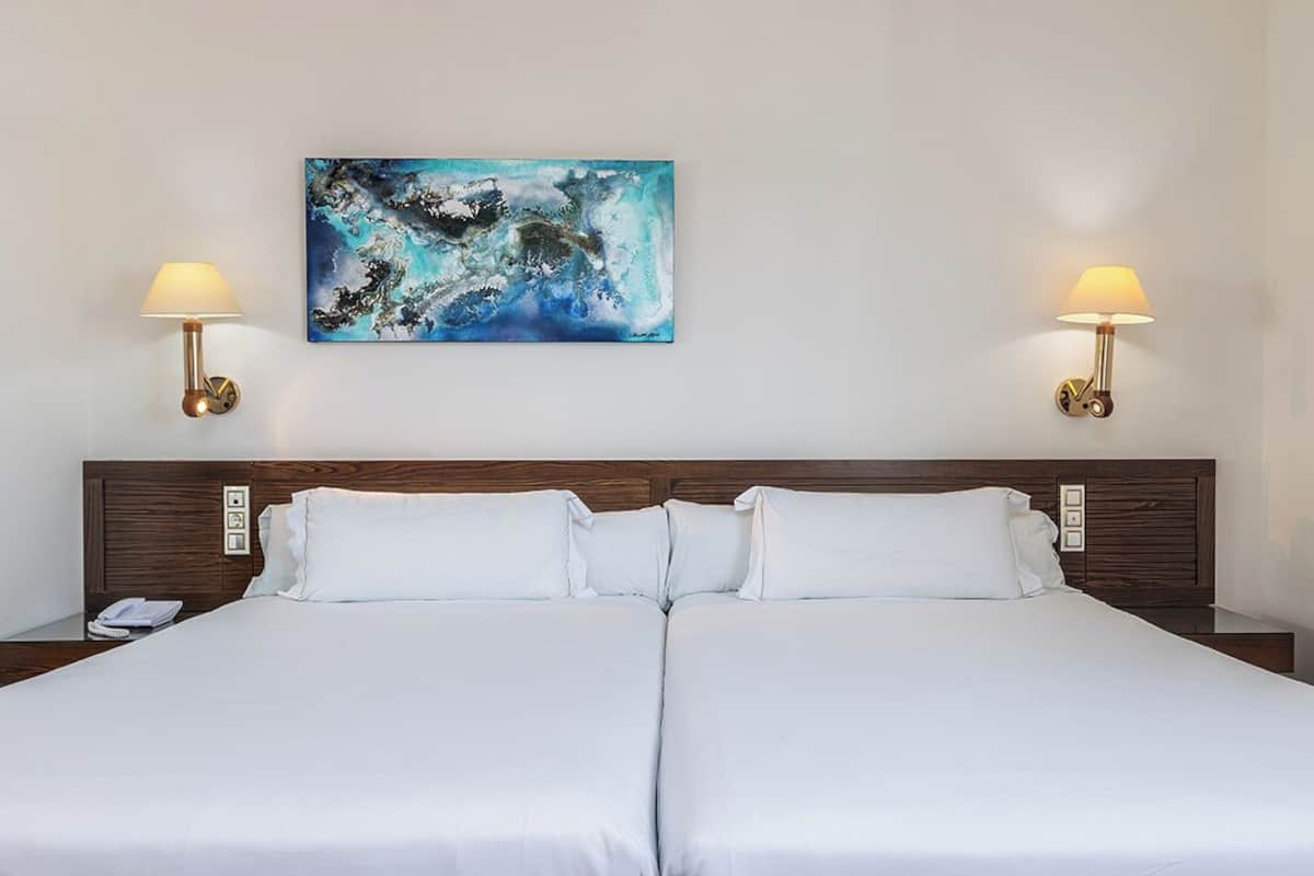 A double bed with white linens, two wall-mounted lamps, and a painting above.