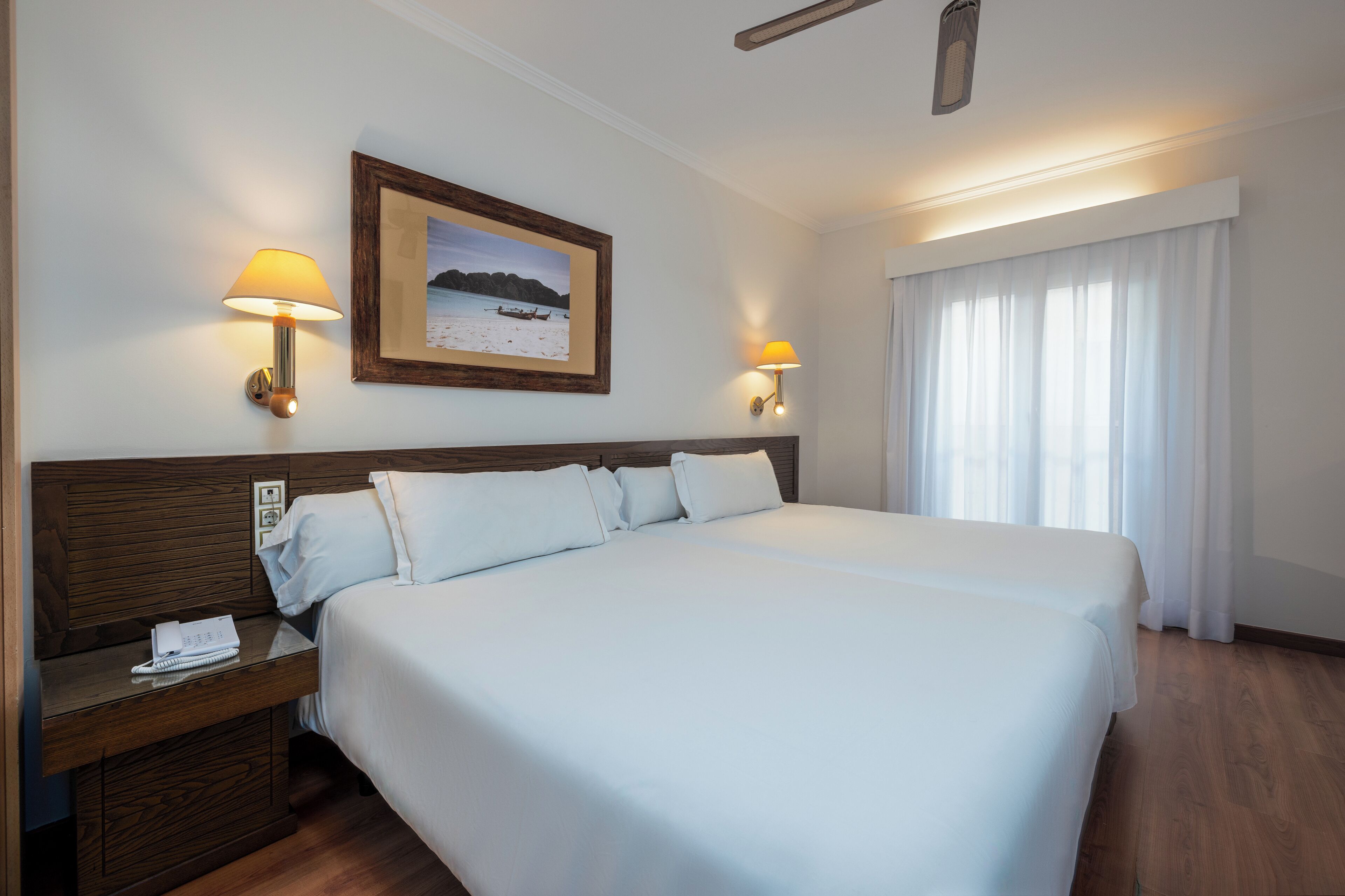 Hotelkamer met een groot bed, twee bedlampen, een nachtkastje met telefoon en notitieboekje, een ingelijst schilderij aan de muur en een raam met gordijnen.