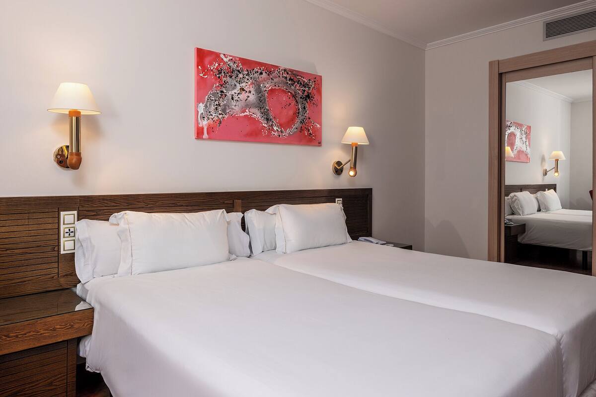 A hotel room with a large bed, two bedside tables, a wooden headboard, a red abstract painting, and two wall-mounted lamps.