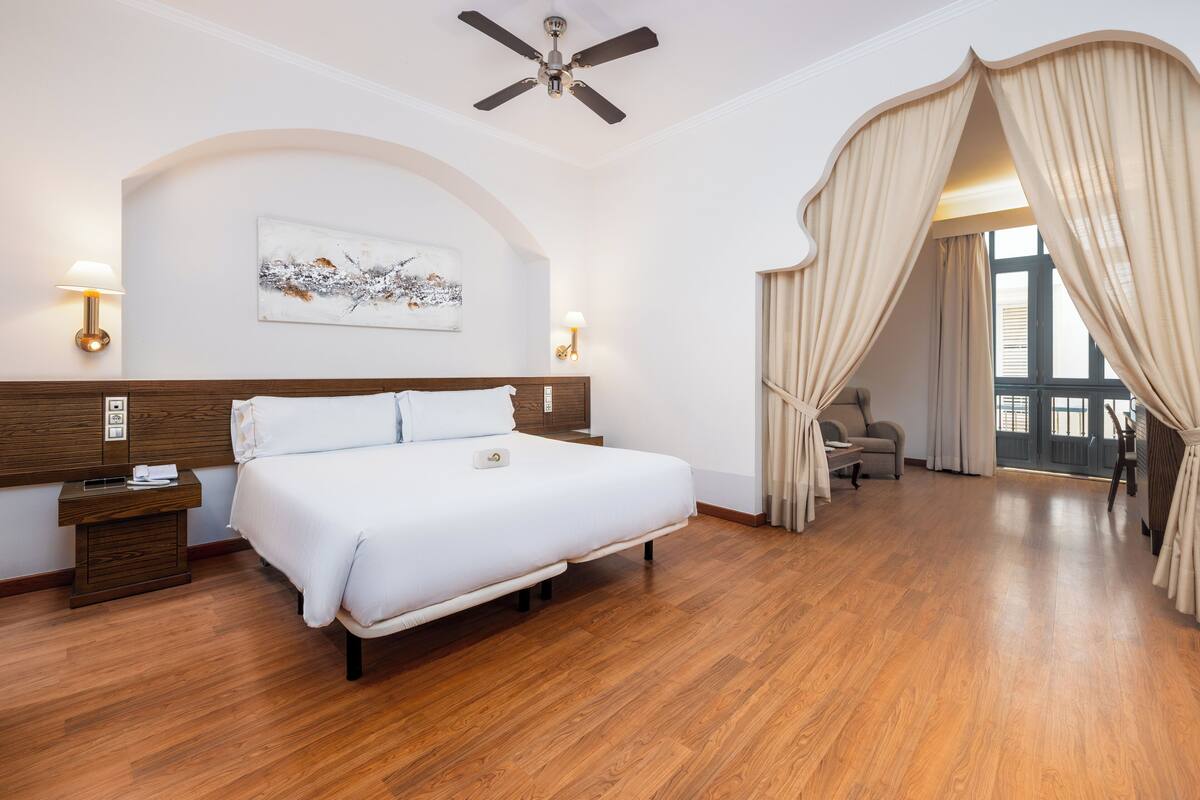A hotel room with a large bed, wooden headboard, a ceiling fan, a painting on the wall, and a view through a window with curtains.
