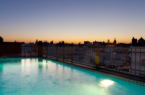 A swimming pool with a geometric tile pattern, surrounded by a metal fence, and a city skyline in the background. - Senator Cadiz Hotel (Cádiz)