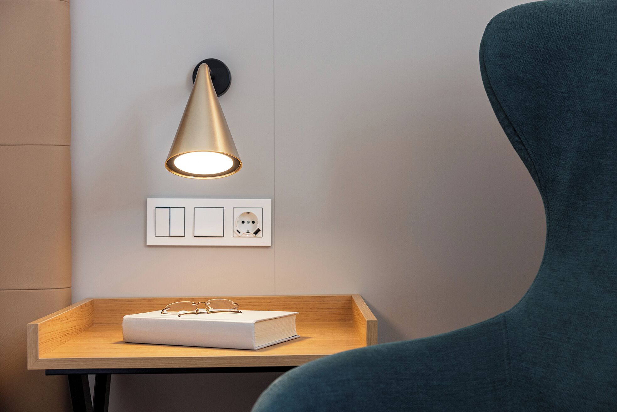 A modern room with a wall-mounted light fixture, a power outlet, and a small wooden table with a book and glasses.
