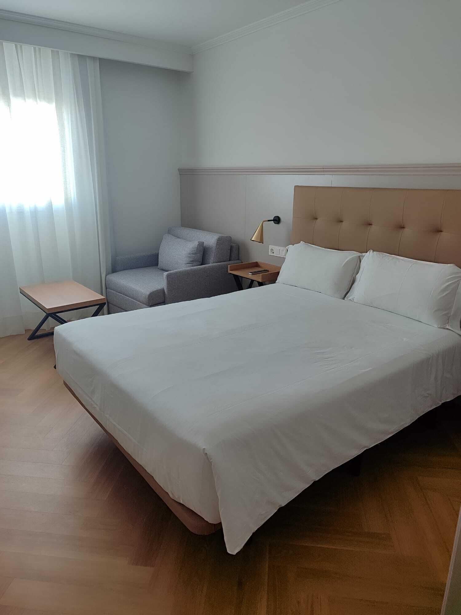 A hotel room with a large bed, a grey armchair, a small table, and a bedside lamp.