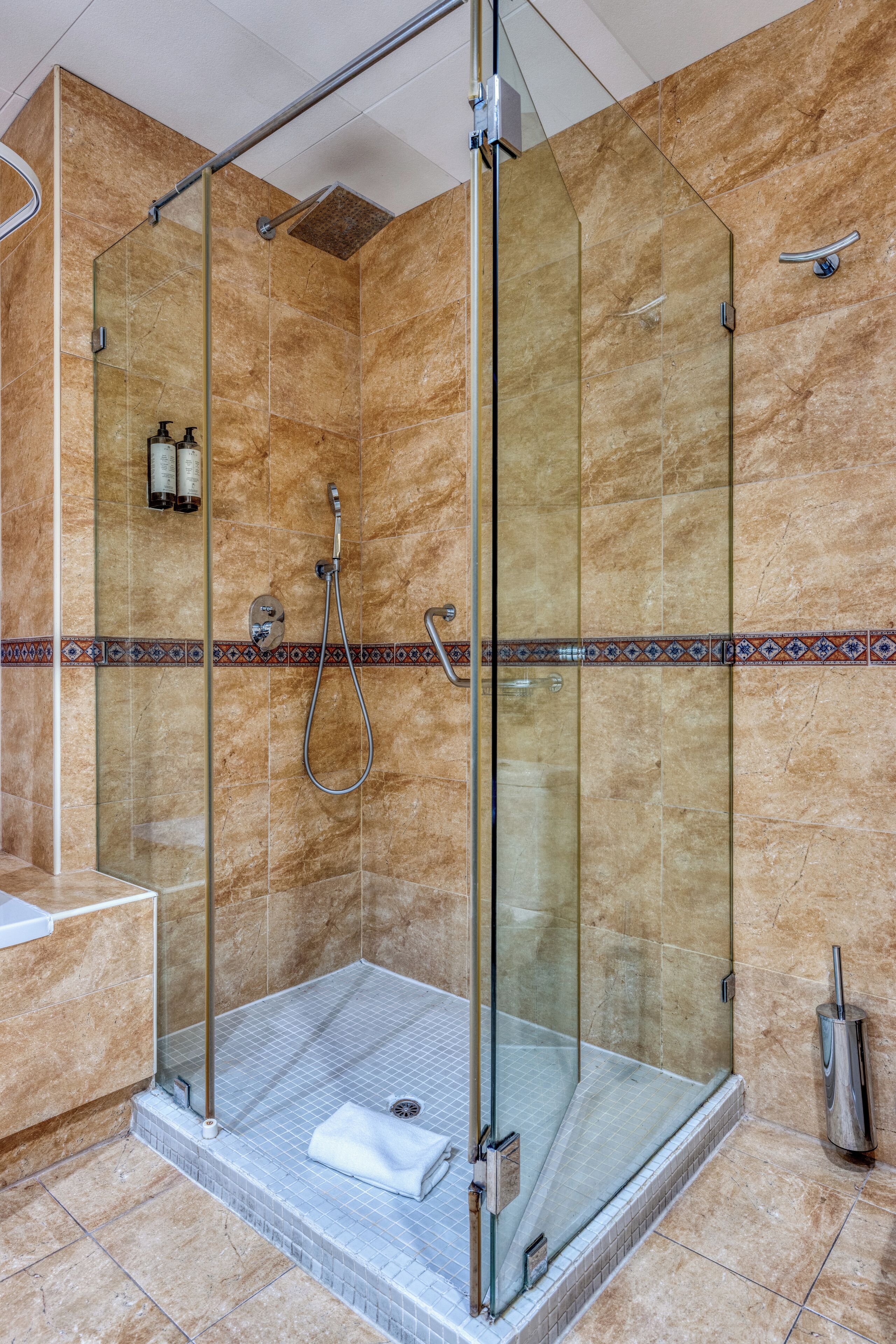 A glass-enclosed shower stall with a tiled floor, a handheld showerhead, and a handheld faucet.