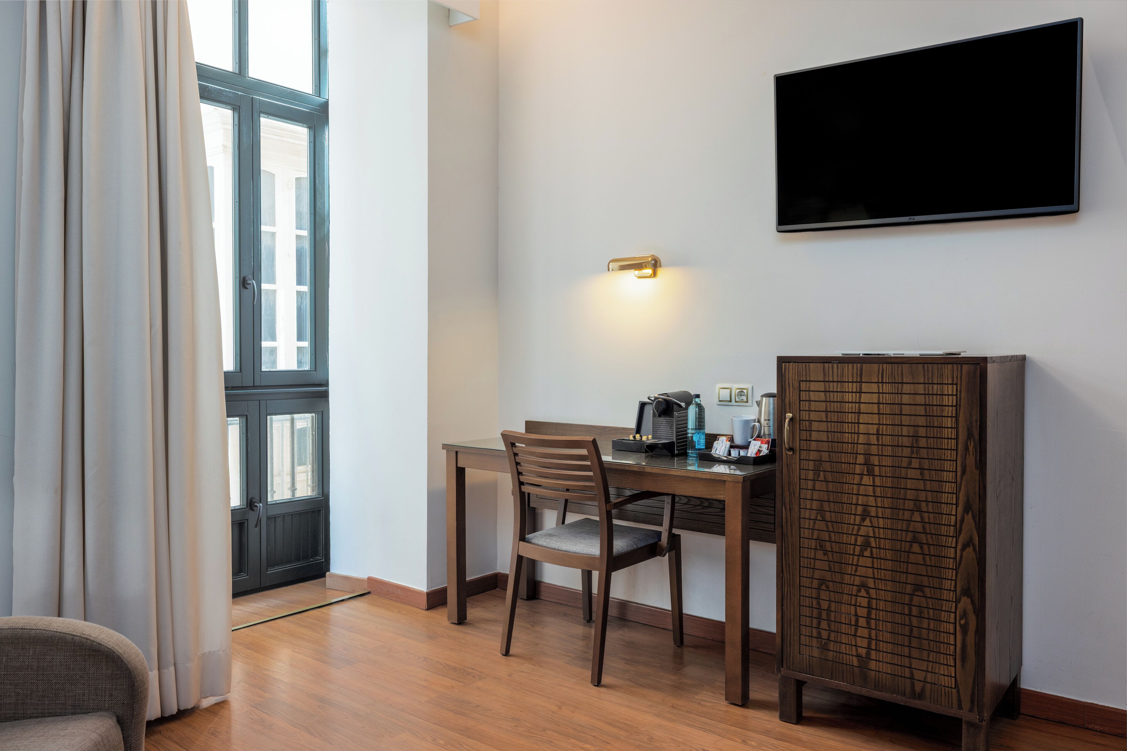 A hotel room with a wooden dining table, a single chair, a television mounted on the wall, and a cabinet with a patterned door.