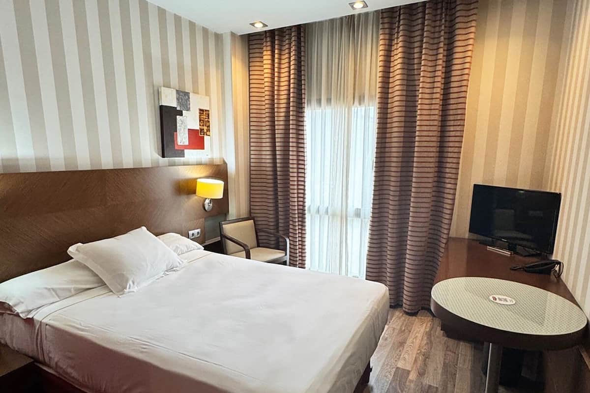 Habitación de hotel con una cama grande, un escritorio con silla, televisor y ventana con cortinas.
