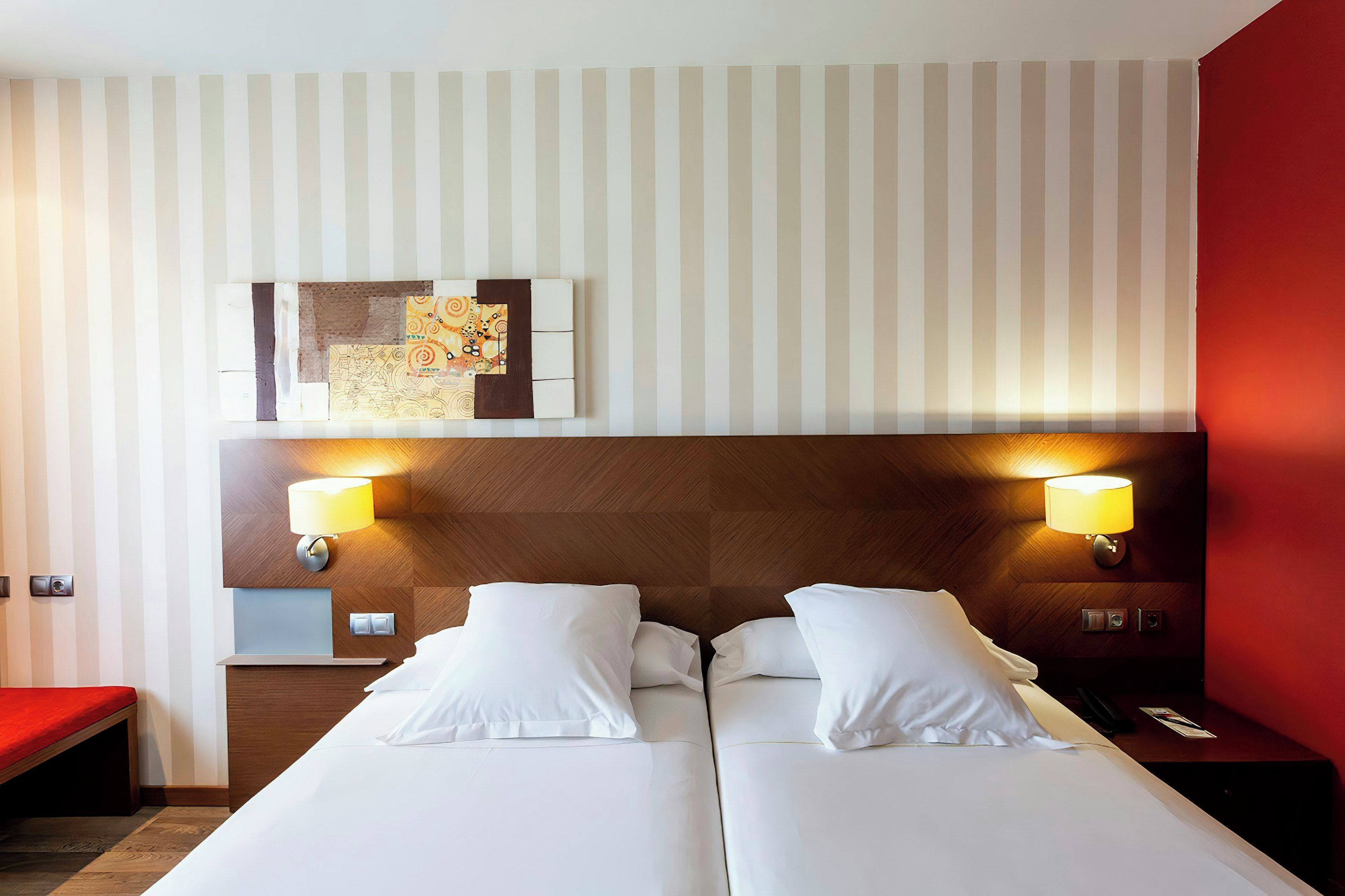 Habitación de hotel con cama doble, dos lámparas de noche, una obra de arte en la pared y una pared de acento roja.