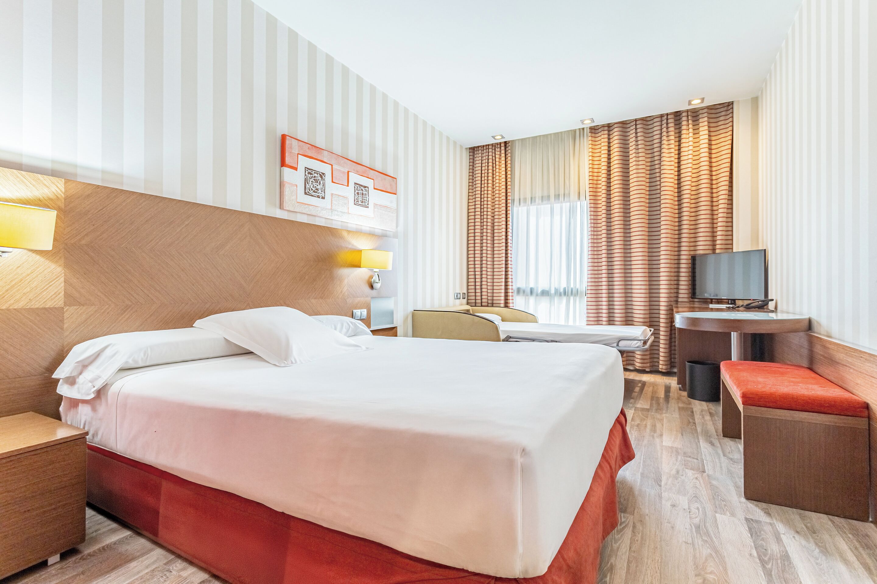 Una camera d'albergo con un letto, comodino, lampada, televisore e una piccola scrivania con sedia.