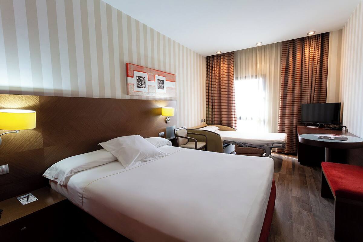 A hotel room with a large bed, bedside tables, a desk, a chair, a TV, and curtains.
