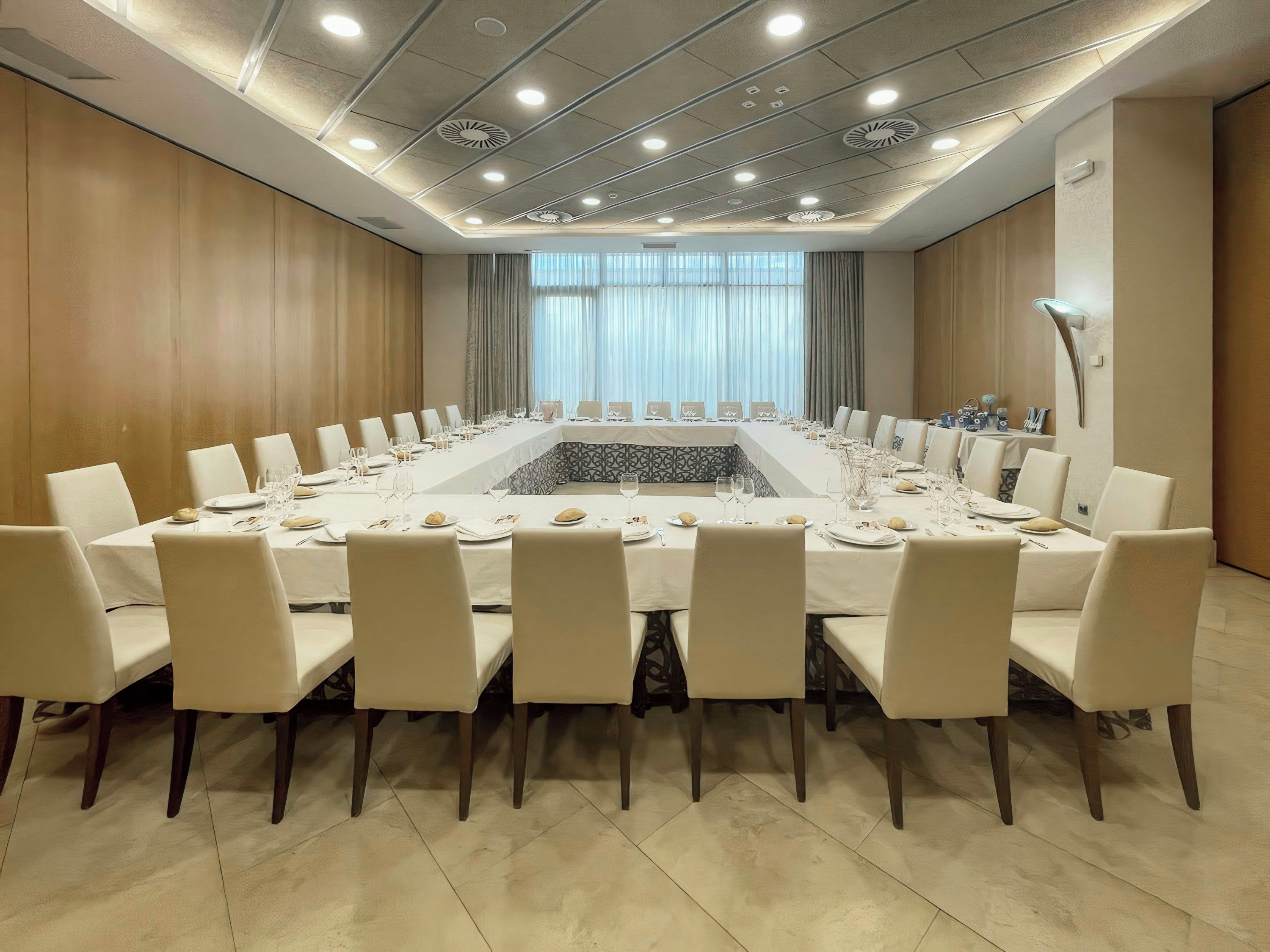 Une salle de conférence dotée d’une longue table préparée pour une réunion, entourée de chaises, avec un plafond au design moderne et de grandes fenêtres.