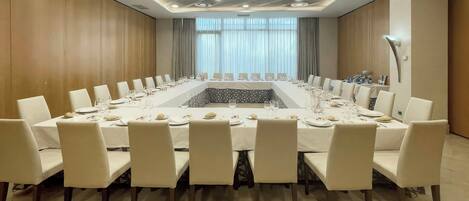 Une salle de conférence dotée d’une longue table préparée pour une réunion, entourée de chaises, avec un plafond au design moderne et de grandes fenêtres.