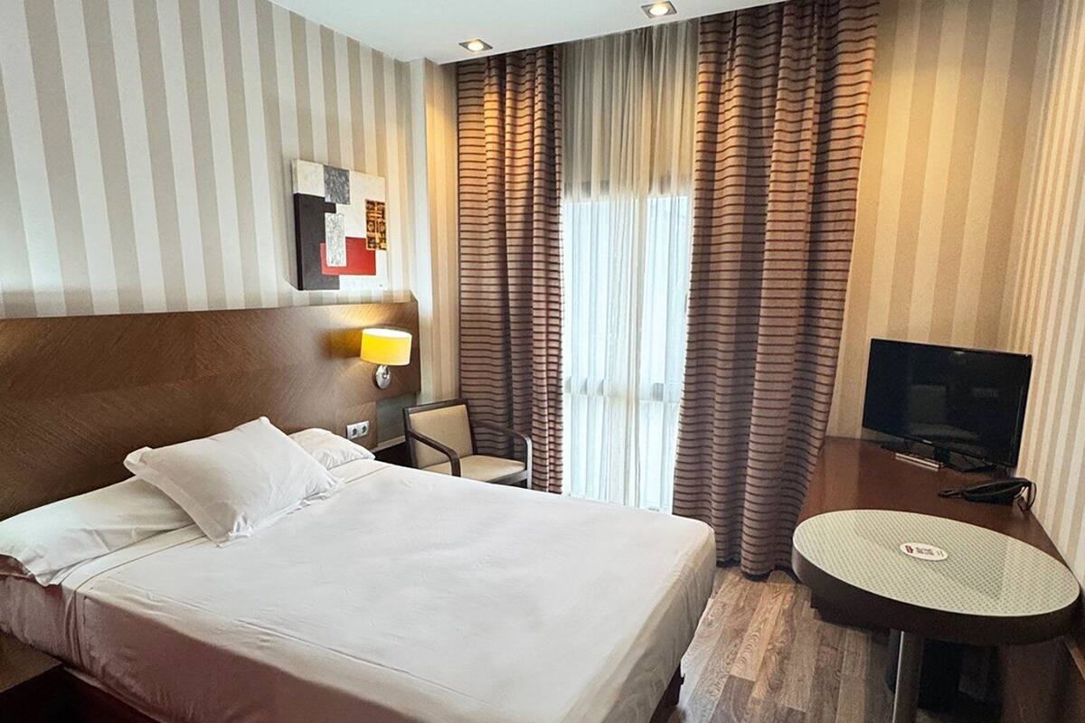 A hotel room with a large bed, a desk with a chair, a television, and a window with curtains.