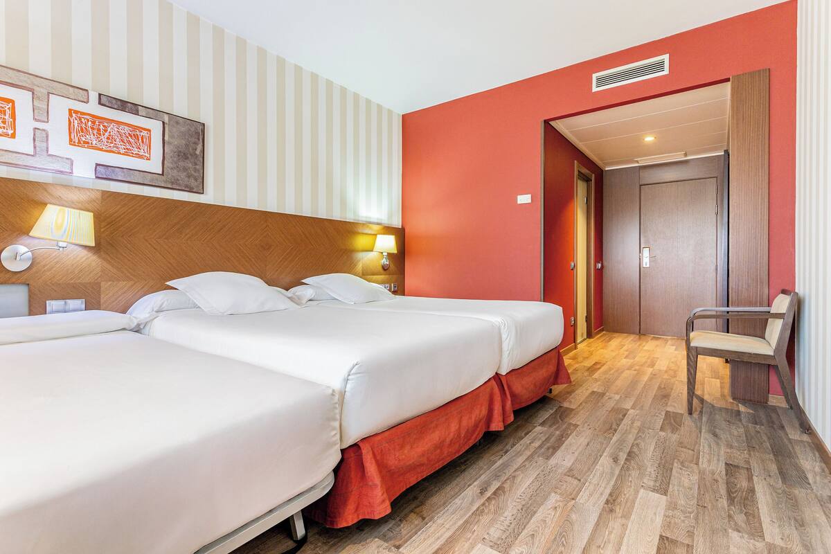 A hotel room with two beds, a wooden headboard, a red accent wall, a chair, and a door leading to another room.