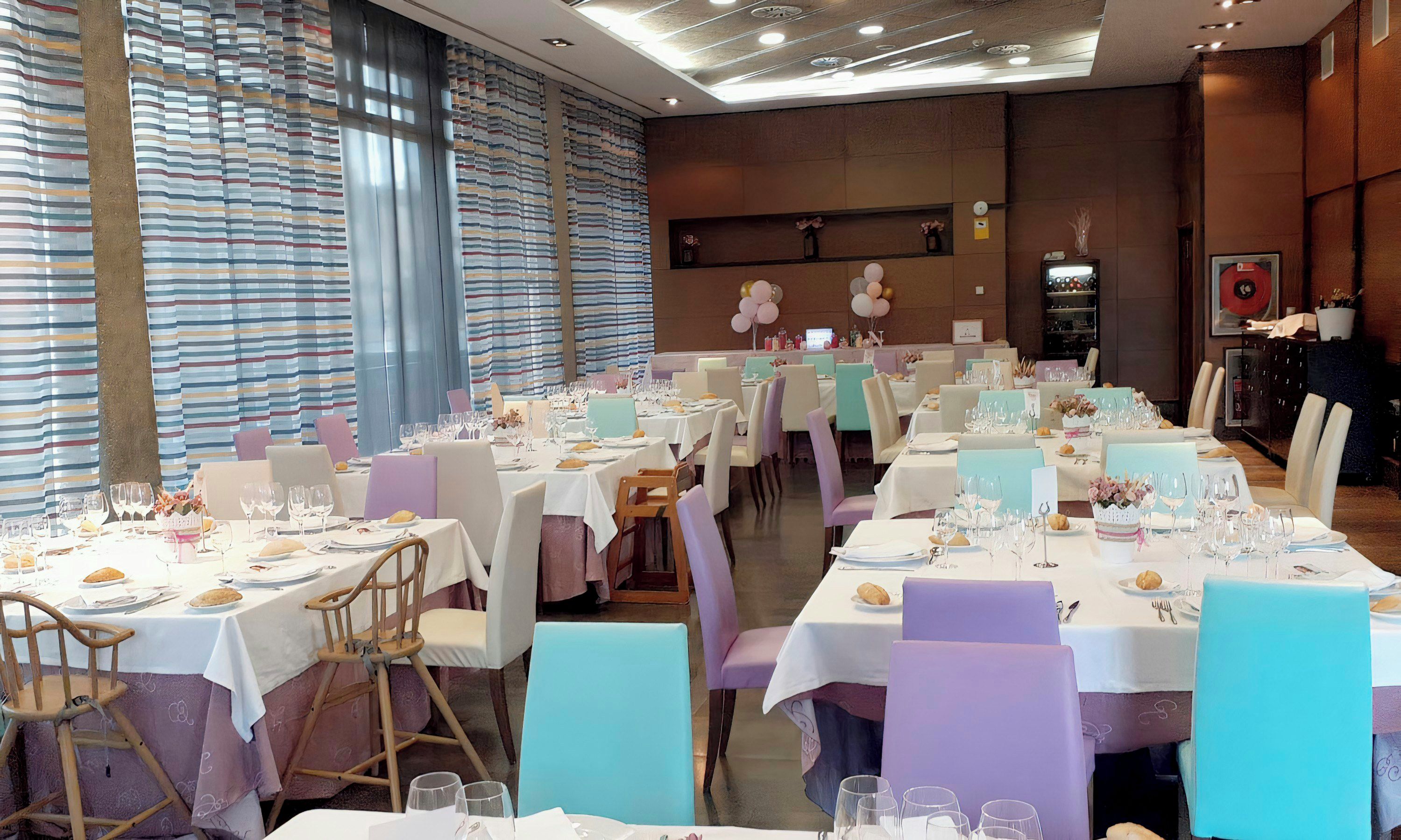 Comedor con varias mesas dispuestas para una comida, sillas alrededor y un amplio ventanal con cortinas.